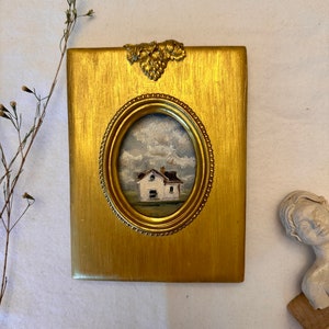 May include: A rectangular gold-toned frame with a textured finish and a decorative grape cluster accent at the top. The frame features an oval center opening containing a small oil painting of a white house under a cloudy sky.
