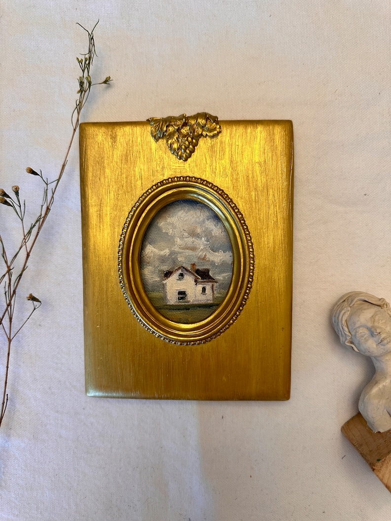 May include: A rectangular gold-toned frame with a textured finish and a decorative grape cluster accent at the top. The frame features an oval center opening containing a small oil painting of a white house under a cloudy sky.