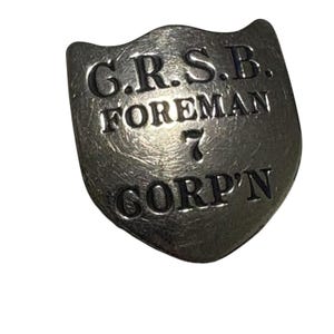 Könnte beinhalten: Ein schildförmiges Metallabzeichen mit dem geprägten Text "G.R.S.B. FOREMAN 7 CORP'N". Das Abzeichen hat ein verwittertes, antikes Aussehen mit einer leicht gebogenen Oberfläche und einer dunklen, metallischen Oberfläche. Der Text ist fett und ohne Serifen.