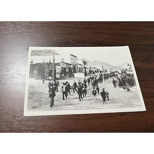 May include: Black and white vintage postcard depicting a parade on Golden Street in Rhyolite, Nevada, in 1906. The image shows a crowd of people walking down the street, with buildings and mountains in the background. An American flag is visible.