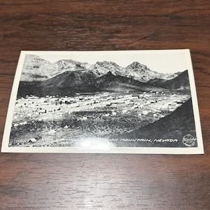 Puede incluir: Postal vintage en blanco y negro que representa un pueblo al pie de una cordillera. La postal rectangular tiene el texto "USH MOUNTAIN, NEVADA" impreso.