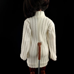 May include: A white ribbed sweater dress with a long sleeve and a turtleneck. The dress is being worn by a doll with brown hair and brown skin.