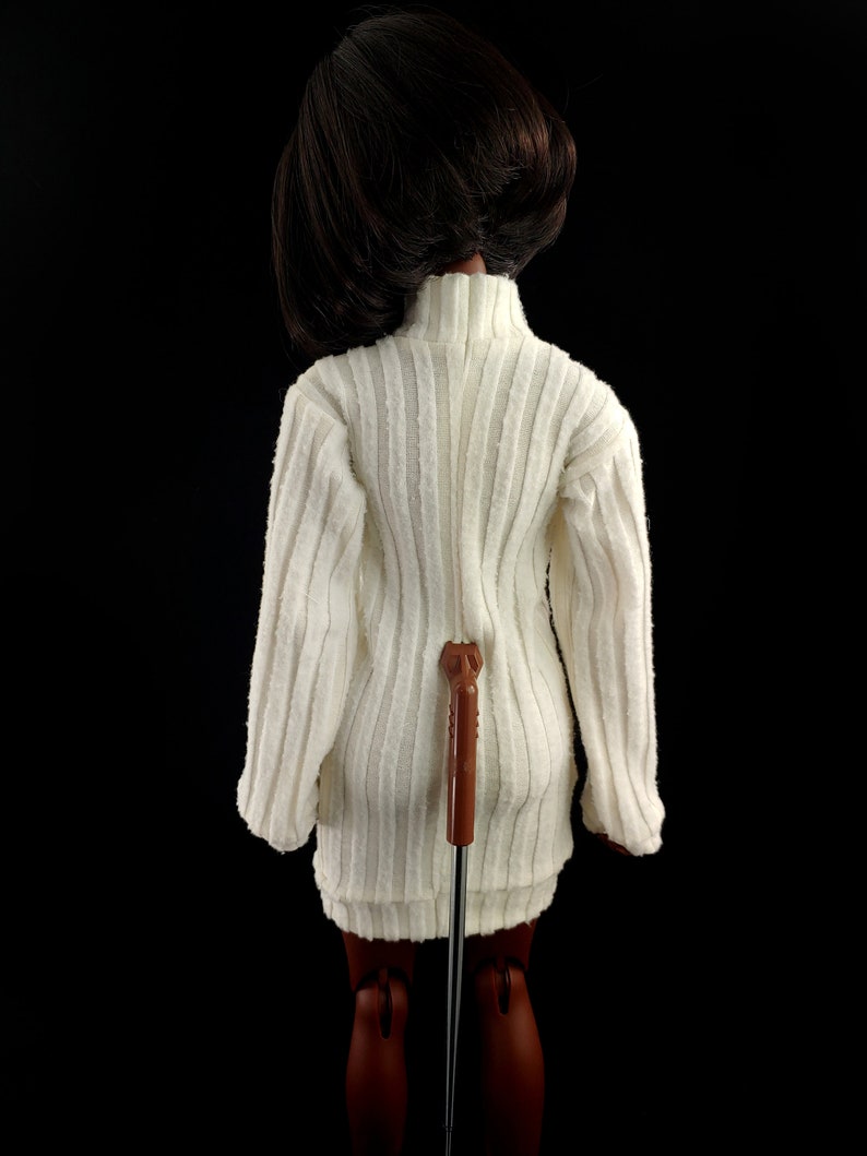 May include: A white ribbed sweater dress with a long sleeve and a turtleneck. The dress is being worn by a doll with brown hair and brown skin.