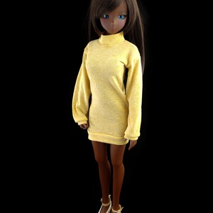 May include: A doll wearing a yellow turtleneck sweater dress with long sleeves. The doll has brown hair and blue eyes.
