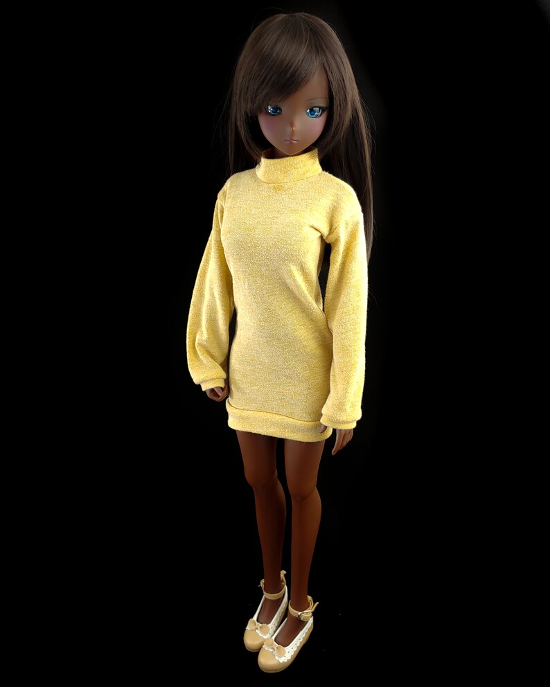 May include: A doll wearing a yellow turtleneck sweater dress with long sleeves. The doll has brown hair and blue eyes.