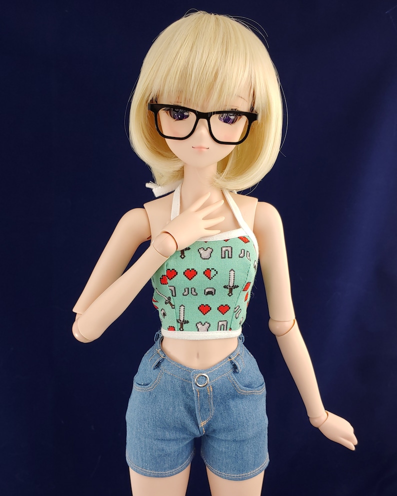 May include: A doll with blonde hair wearing black glasses, a green and white halter top with a pixelated design, and blue denim shorts.