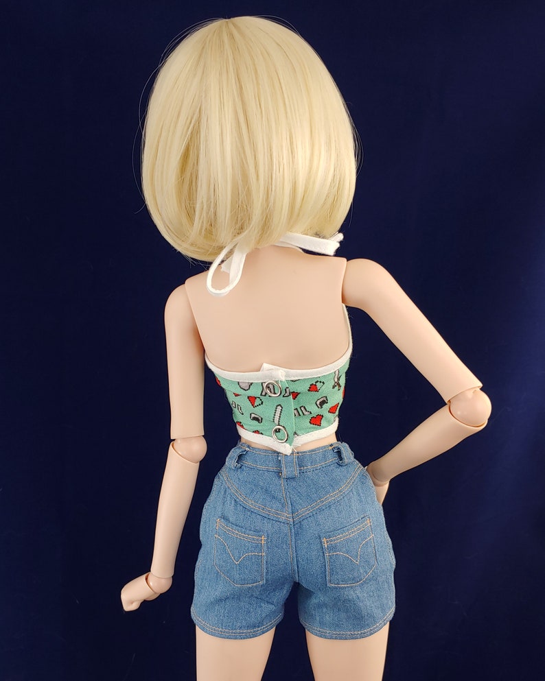 May include: A doll wearing a green and white halter top with a red and white pattern and blue denim shorts.