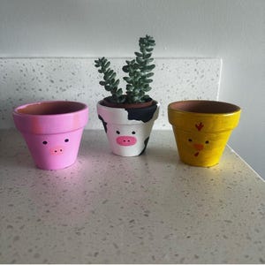 May include: Three small terracotta plant pots painted as animals. The left pot is pink with a pig face. The center pot is white with black spots, a pink snout, and a green succulent. The right pot is yellow with a chick face and red beak.