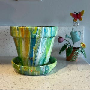 May include: A ceramic plant pot and matching saucer featuring a marbled paint pour design in shades of green, blue, yellow, and white. Beside it stands a decorative metal garden stake with a light blue watering can and a yellow butterfly.