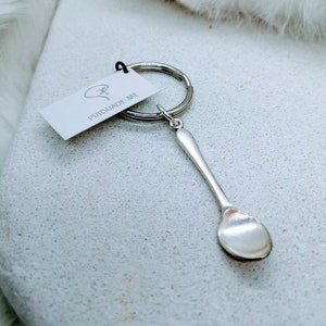 May include: A silver-colored spoon-shaped keychain with a small tag attached. The tag reads "PERSUADE ME" in black text. The keychain is on a silver metal ring.
