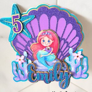 May include: A mermaid-themed cake topper with a glittery blue starfish and the number 5. The topper features a mermaid, a seashell, and the name "Emily" in blue glitter. Coral and flowers add detail.