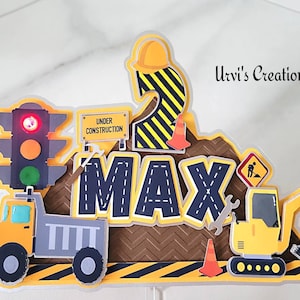 May include: A yellow and black construction themed cake topper with the name "Max" and the number "2" on it. The topper includes a dump truck, an excavator, traffic lights, and a sign that says "Under Construction".