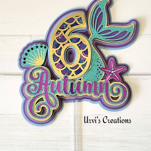 May include: A mermaid-themed cake topper with a gold glitter number 6, a purple and teal mermaid tail, and the name "Autumn" in purple glitter. The topper is on a white wooden background.