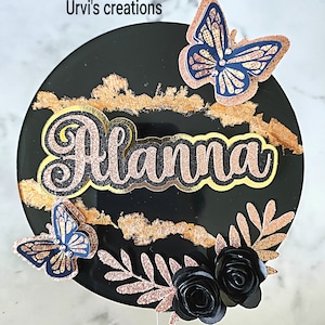 May include: A circular black cake topper featuring the name Alanna in glittery rose gold script. The design includes two navy and rose gold butterflies, two black paper roses, rose gold glittery foliage, and text reading Urvi's creations.