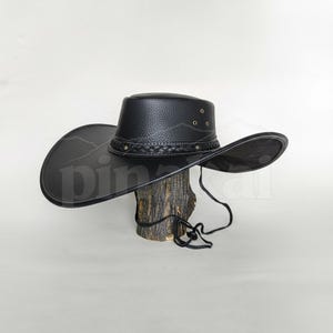 May include: A black cowboy hat with a wide brim and a braided band. The hat is made of a textured material and has a chin strap. The hat is displayed on a wooden stand.