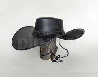 May include: A black cowboy hat with a wide brim and a braided band. The hat is made of a textured material and has a chin strap. The hat is displayed on a wooden stand.