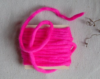 Puede incluir: Un carrete de hilo de lana de color rosa brillante, enrollado firmemente y listo para proyectos de manualidades.