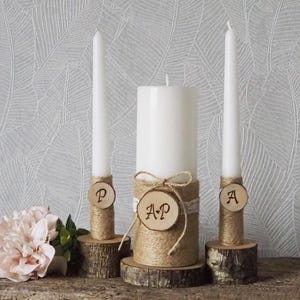 May include: Three white candles of varying heights, decorated for a wedding. The candles are on wooden bases and wrapped with twine. Wooden discs with initials are attached. A pink flower and greenery are in the foreground.