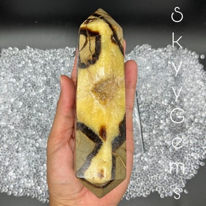 May include: A large, light brown and dark brown banded agate crystal point, also known as a Botswana agate, is held in a hand. The crystal point is approximately 15 cm long and has a smooth, polished surface.