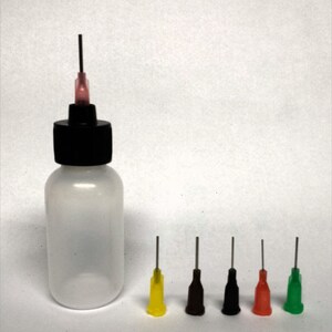May include: A clear plastic bottle with a black cap and a dispensing needle. Five additional dispensing needles with colored tips in yellow, brown, black, red, and green are arranged to the right of the bottle. The needles are for dispensing liquids.