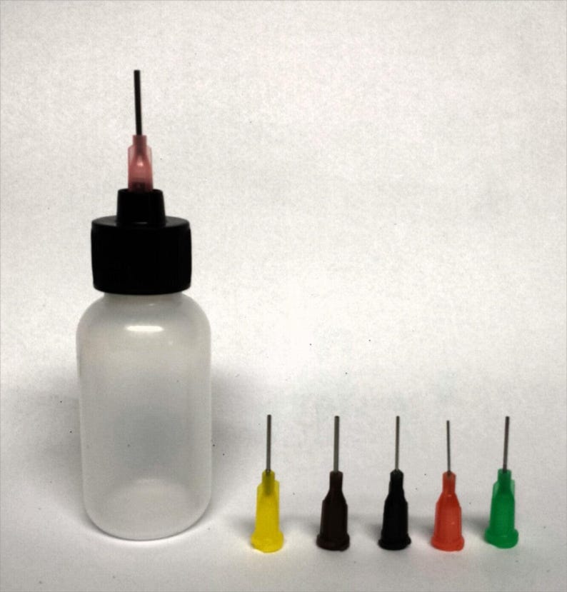 May include: A clear plastic bottle with a black cap and a dispensing needle. Five additional dispensing needles with colored tips in yellow, brown, black, red, and green are arranged to the right of the bottle. The needles are for dispensing liquids.