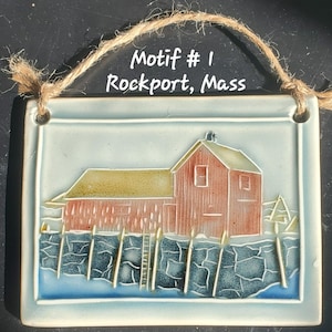 Puede incluir: Azulejo cerámico rectangular con una escena costera. El azulejo presenta un edificio rojo con techo amarillo, un océano azul y un muelle de piedra. El texto "Motif #1 Rockport, Mass" está escrito en el azulejo. Una cuerda de yute está adjunta para colgar.