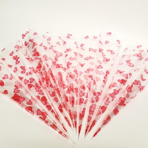 May include: Several transparent cellophane cones are fanned out, each adorned with scattered red heart shapes. The cones are arranged in a radial pattern, with the tips pointing towards the center. The background is a plain white surface.