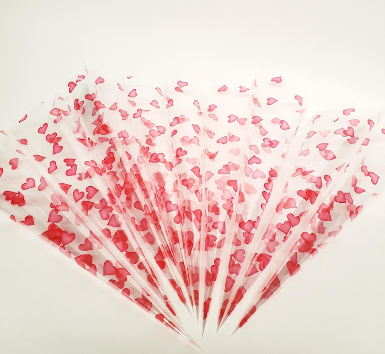May include: Several transparent cellophane cones are fanned out, each adorned with scattered red heart shapes. The cones are arranged in a radial pattern, with the tips pointing towards the center. The background is a plain white surface.