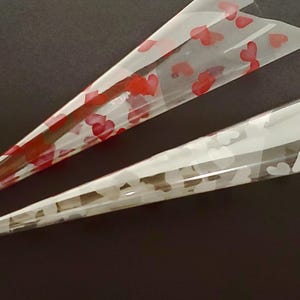 May include: Two transparent, cone-shaped henna applicators. One is decorated with red heart patterns, the other with white heart patterns. The cones are on a dark surface, with the tips pointing towards the bottom left.