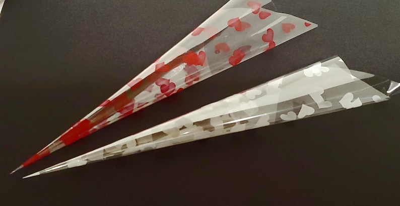 May include: Two transparent, cone-shaped henna applicators. One is decorated with red heart patterns, the other with white heart patterns. The cones are on a dark surface, with the tips pointing towards the bottom left.
