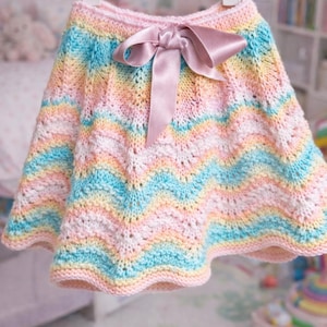 May include: A pastel-colored, knitted skirt with a chevron pattern. The skirt features a pink waistband and a matching pink satin bow. The skirt is displayed on a white hanger, suggesting it's a children's garment.