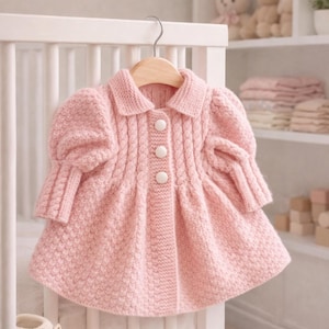 May include: A light pink knitted baby coat with a collared neckline and three white buttons. The coat has a textured pattern and puffy sleeves. A pair of white knitted baby booties are in the foreground. The coat is hanging on a wooden hanger.