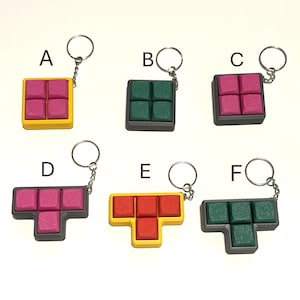 May include: Six keychains featuring Tetris block designs. Each keychain has a silver ring and chain. The blocks are in various colors, including pink, green, and red, with yellow and gray frames. The keychains are arranged on a white surface.