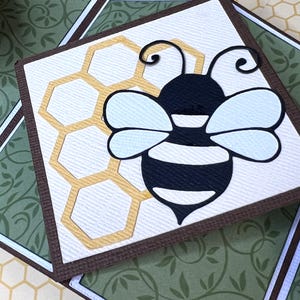 May include: A square card with a brown border features a black and white bee with light blue wings. A yellow honeycomb design is visible next to the bee. The background has a green floral pattern.