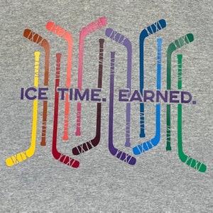 May include: A gray sweatshirt featuring a design of colorful hockey sticks. The sticks are arranged vertically and in a row. The text "ICE TIME. EARNED." is printed in purple below the hockey sticks.
