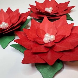 May include: Three red paper poinsettia flowers with white pearl embellishments. The flowers have green paper leaves.