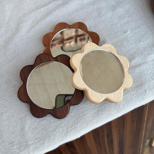 May include: Three small, flower-shaped mirrors. One is made of light wood, one of medium-toned wood, and one of dark wood. Each mirror has a circular reflective surface. The mirrors are arranged on a light-colored, textured surface.