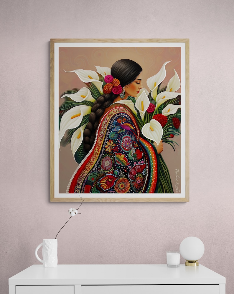 May include: A woman in a colourful embroidered dress holds a bouquet of white calla lilies. The flowers are arranged in a fan shape, with the woman's hand visible at the bottom of the bouquet. The woman's hair is long and dark, and she is wearing a flower in her hair. The background is a light brown colour.