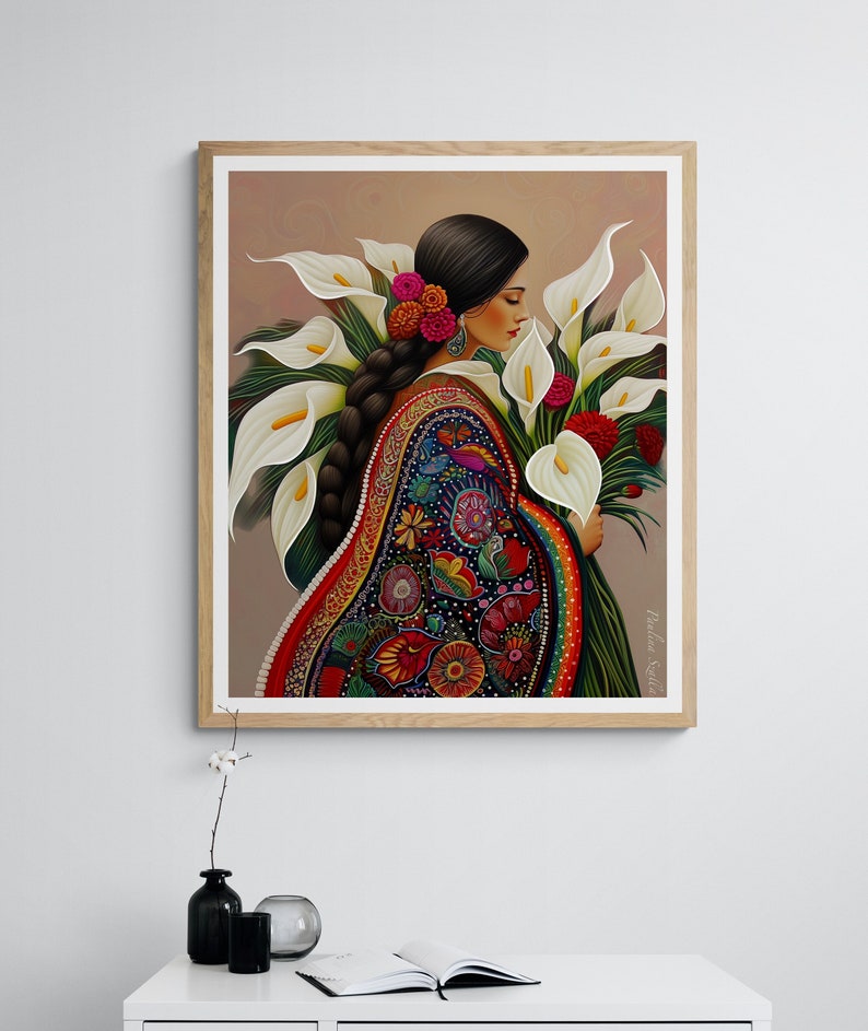 May include: A framed illustration of a woman in a colourful traditional Mexican dress holding a bouquet of white calla lilies. The woman has long dark hair and is wearing a beaded necklace. The background is a light brown colour.