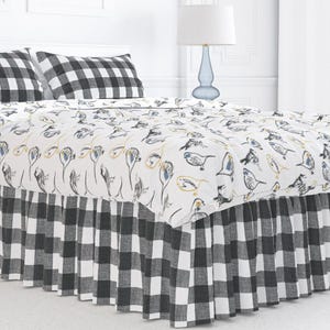 May include: A bed with a white comforter featuring a bird and floral pattern, paired with black and white checkered pillowcases and a matching bed skirt. The bed is made up with a white headboard and a blue lamp.
