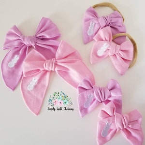May include: Set of four pink and purple fabric hair bows with white embroidered bunny designs. The bows are tied with a knot in the center and are attached to elastic headbands. The text "Simply Quite Charming" is visible in the image.