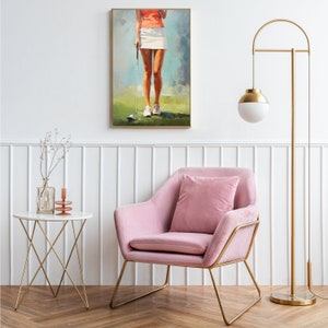 May include: A pink velvet armchair with gold metal legs sits in front of a white wall with a gold floor lamp and a framed painting of a woman playing golf. A white marble side table with a gold metal frame sits in the foreground.