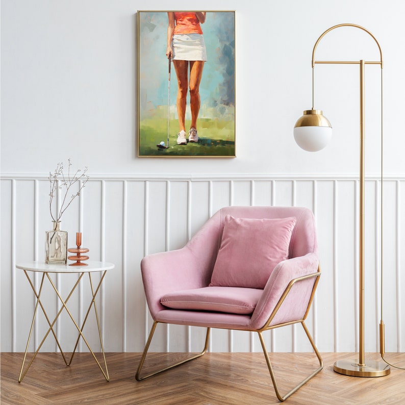 May include: A pink velvet armchair with gold metal legs sits in front of a white wall with a gold floor lamp and a framed painting of a woman playing golf. A white marble side table with a gold metal frame sits in the foreground.