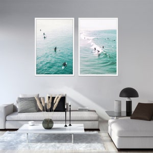 May include: Two framed wall art prints depicting surfers in the ocean. One print shows surfers paddling in calm water, the other shows surfers riding waves. The prints have white frames and are displayed above a modern living room setting.
