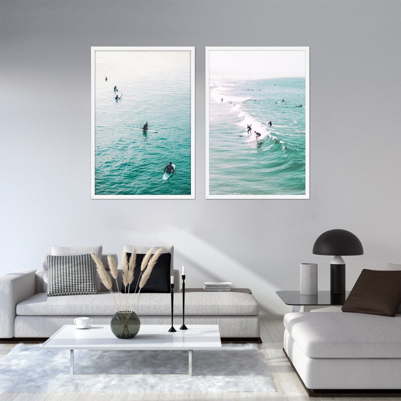 May include: Two framed wall art prints depicting surfers in the ocean. One print shows surfers paddling in calm water, the other shows surfers riding waves. The prints have white frames and are displayed above a modern living room setting.