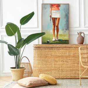 May include: A woven wicker storage trunk with a light brown finish. The trunk is decorated with a painting of a woman playing golf. The painting is framed in a light wood frame. The trunk is sitting on a rug with a green and brown pattern.