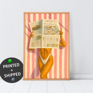 May include: A framed art print featuring a woman in a white swimsuit, holding a newspaper with the headline "OFF DUTY Oh, to Dress Up." The background has pink and white stripes. The artwork has a warm, summery vibe.