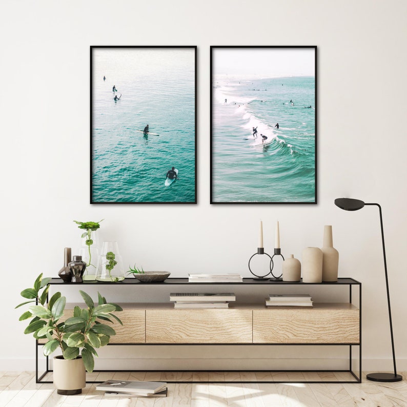 May include: Two framed art prints depict surfers in the ocean. One shows surfers paddling on calm water, the other shows surfers riding waves. The water is a vibrant turquoise color, and the frames are black.