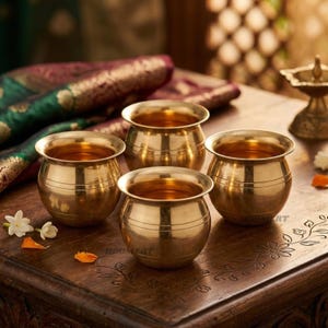 May include: A set of four small, polished brass pots with rounded bodies and wide, flared rims. Each pot features subtle horizontal line detailing around the upper section. The metallic vessels have a warm, reflective gold-toned finish.