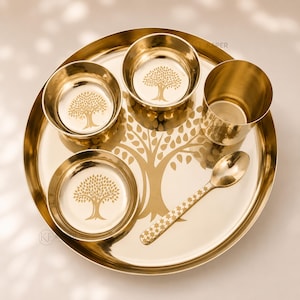 May include: A polished gold-toned metal dinnerware set consisting of a large round tray, two small bowls, one tall tumbler, and a spoon. Each piece features a laser-etched tree design, with the tray displaying a large central tree motif.